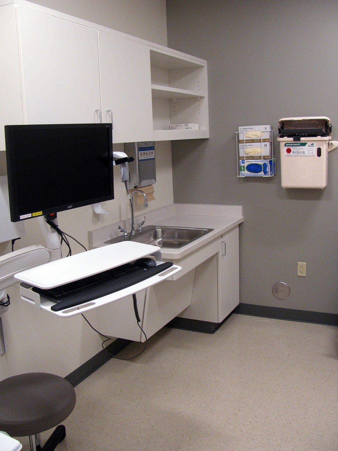 Examination room with a desk, sink, and computer. White cabinets and gray walls.