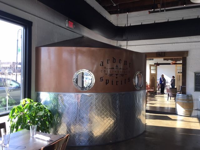 Copper and steel distillery vat at Arden Spirits, with patrons and window view.