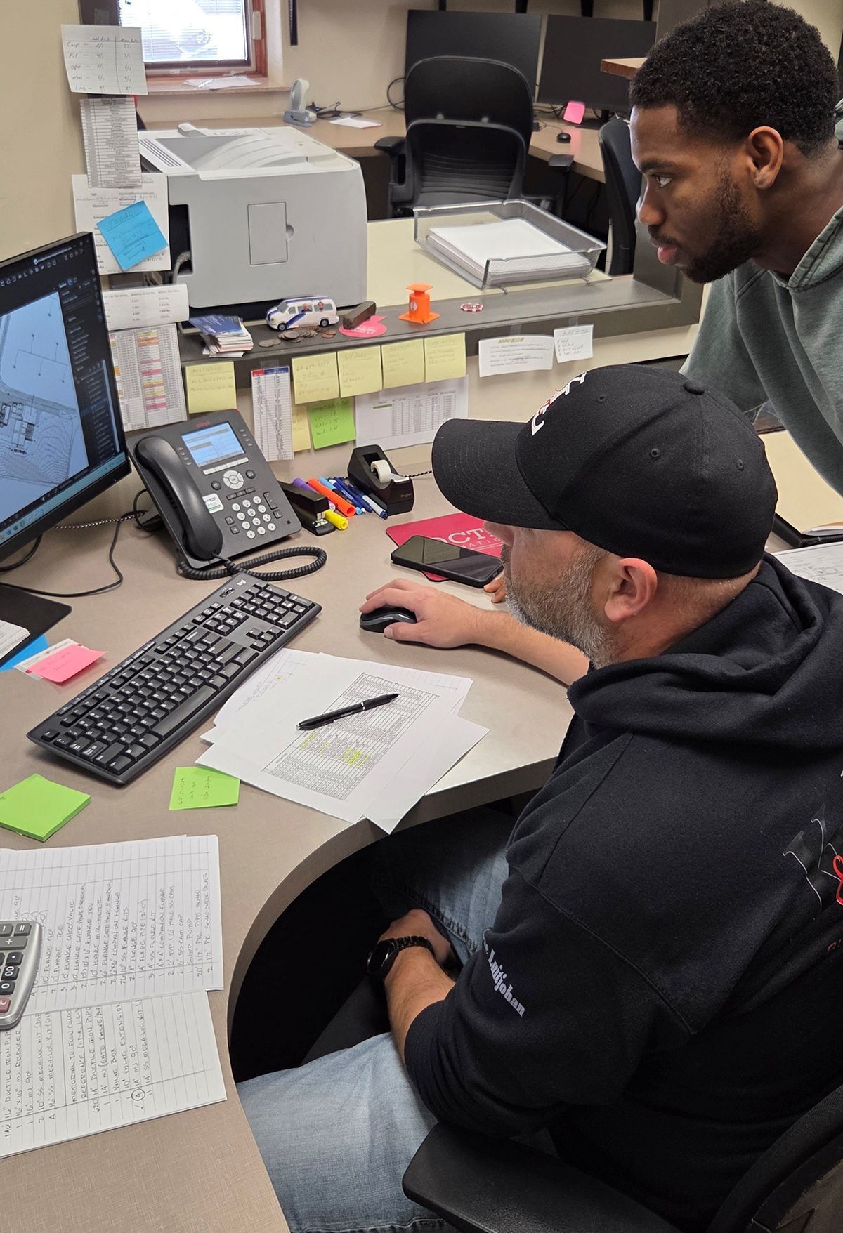 Man and woman in red shirts near a Kubota KX052 excavator. Woman sits in cab, man stands with hand on hip. Outdoors.