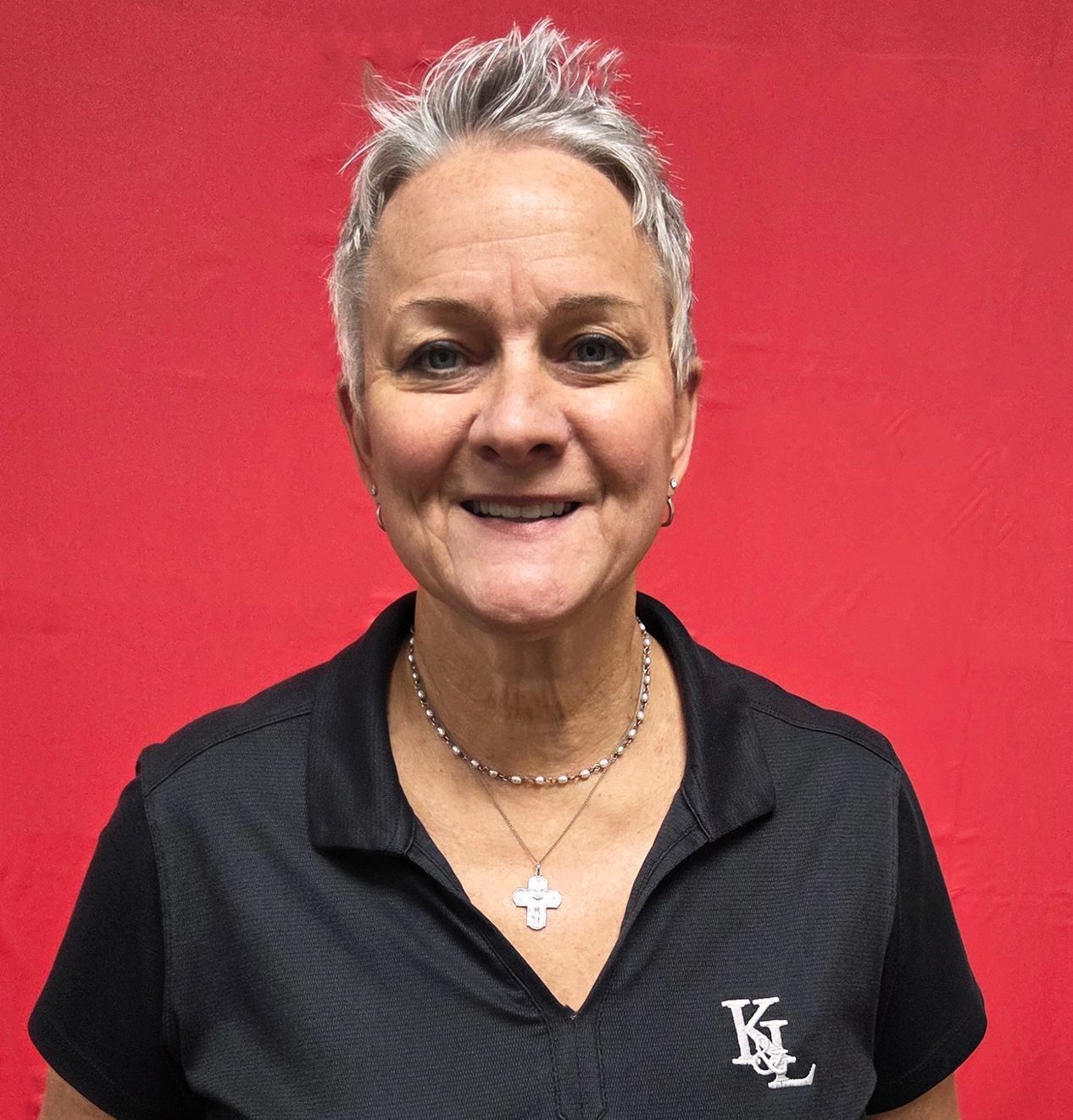 Woman with short gray hair, smiling, wearing a black shirt and necklace, standing in front of a brick wall.
