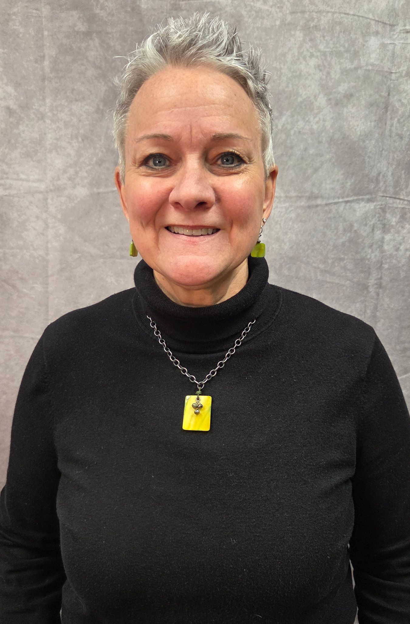 Woman with short gray hair, smiling, wearing a black shirt and necklace, standing in front of a brick wall.