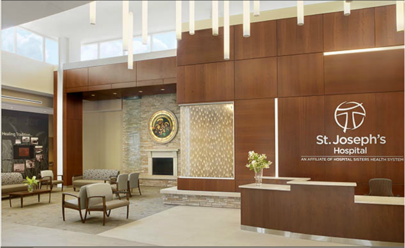Hospital lobby with reception desk, stone fireplace, and seating area. St. Joseph's Hospital logo on wood paneling.