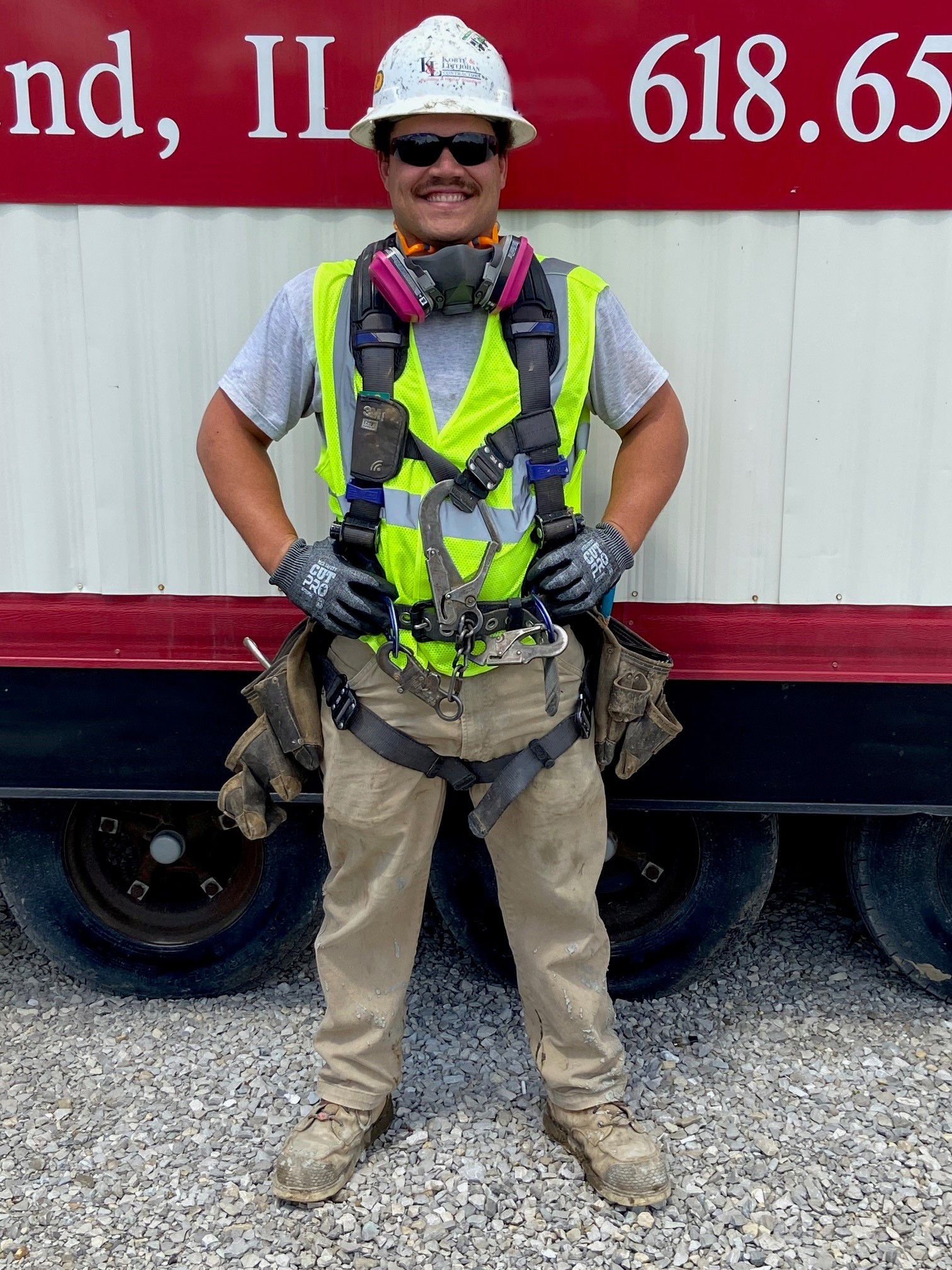 Construction worker in safety gear stands in front of a red building with a phone number on it.