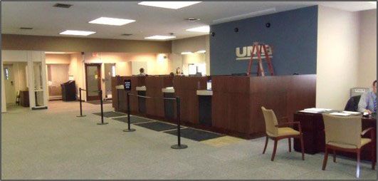 Bank interior with tellers behind a dark wood counter, poinsettias, a person in a uniform, and a waiting area.