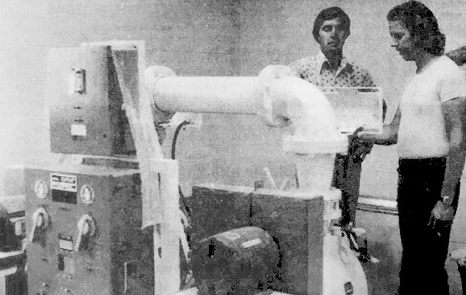 Two men holding an object, seated in front of a company sign that reads