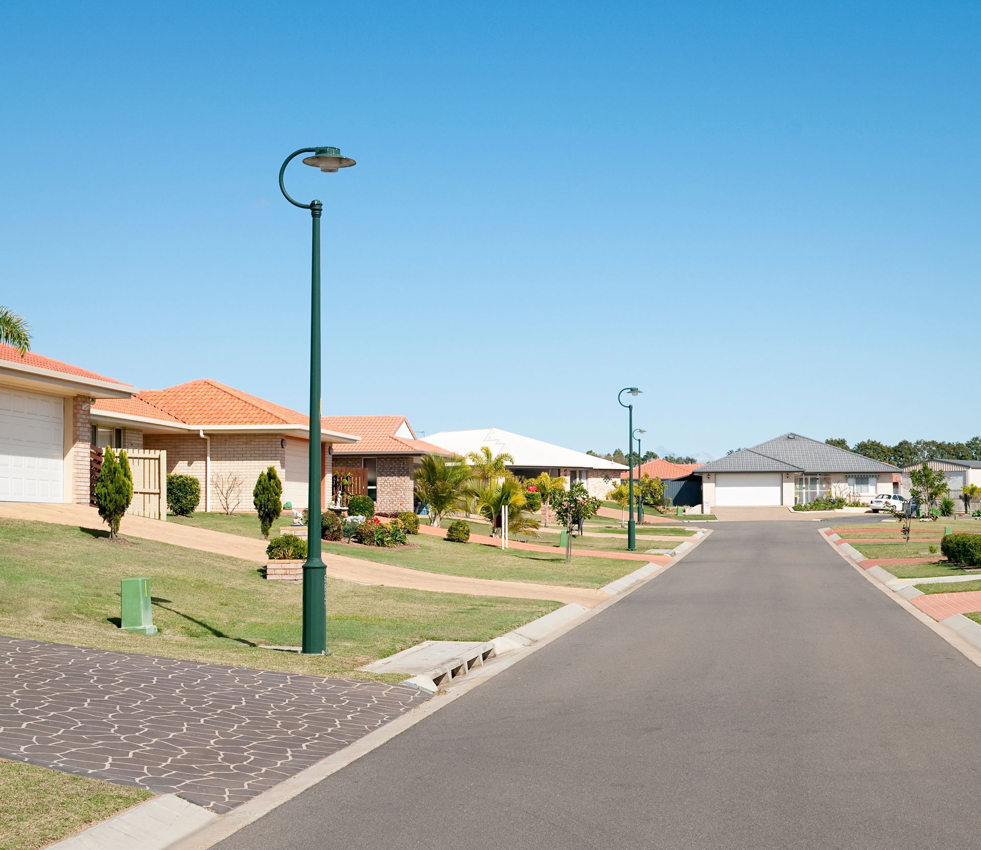Quiet residential street with houses, green lawns, and streetlights under a clear blue sky.