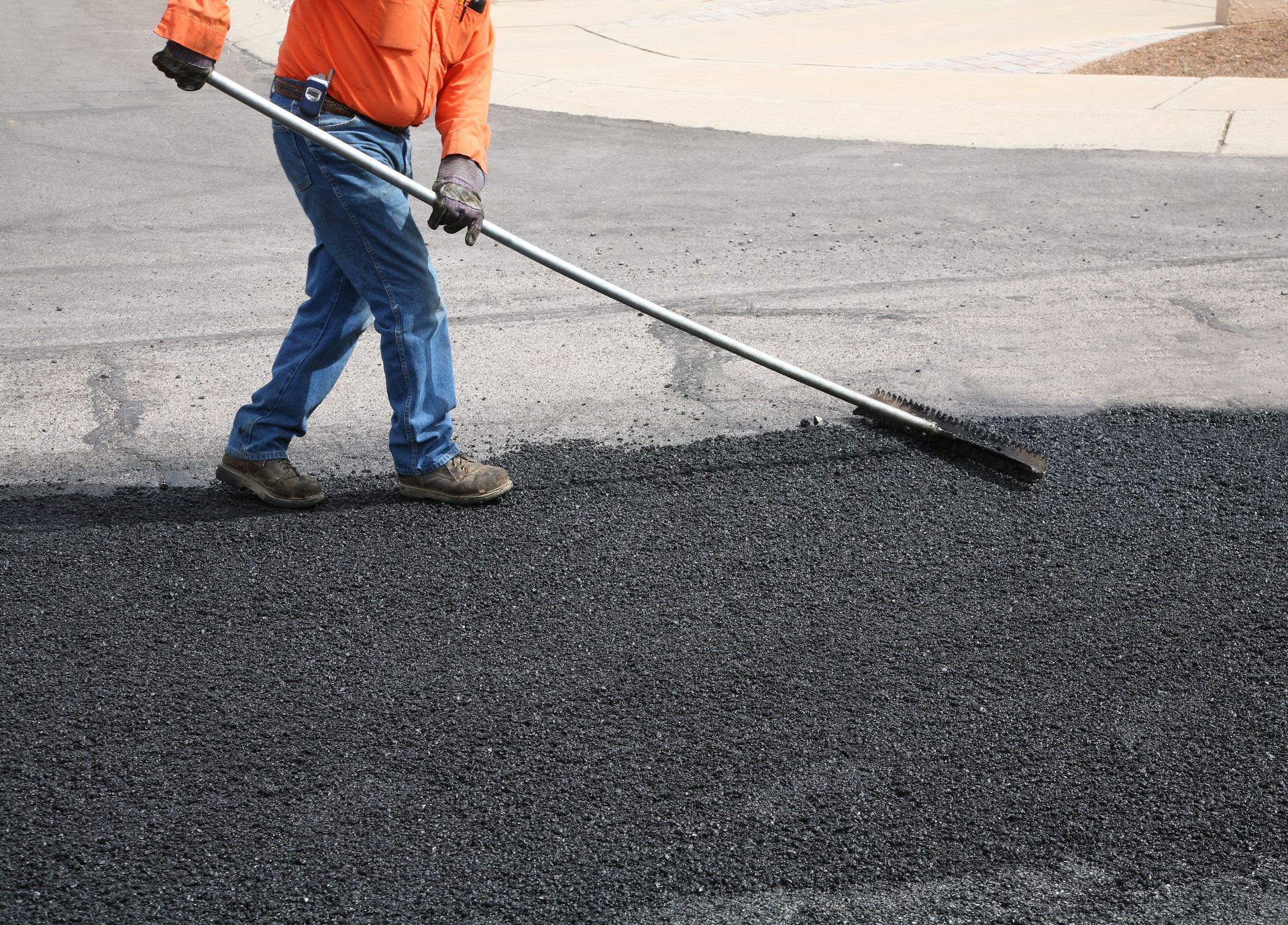 Person in orange shirt raking asphalt on a road.