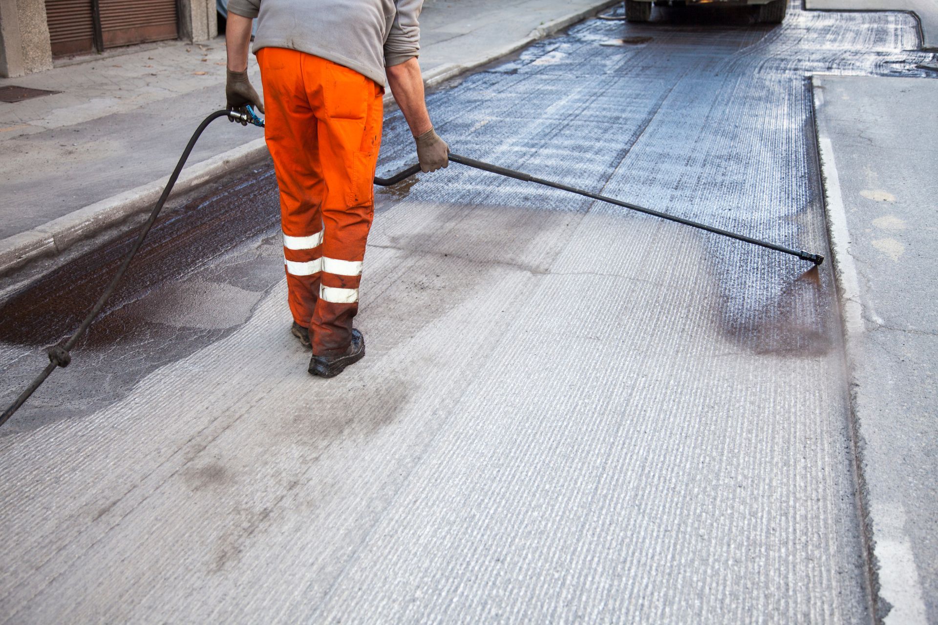 Worker spraying asphalt sealant on road, wearing orange pants and safety boots.