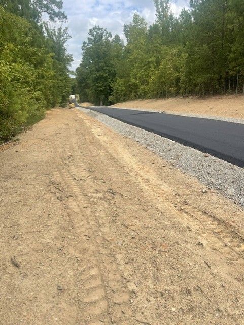 New paved road with gravel shoulder and dirt path alongside, flanked by trees.