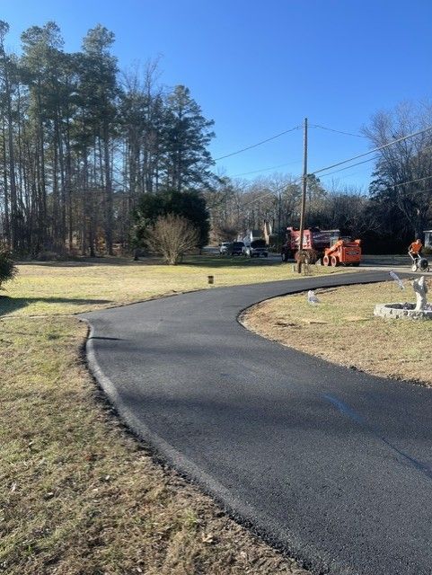 Curving black asphalt driveway with a dark border, leading to a grassy area under a blue sky.