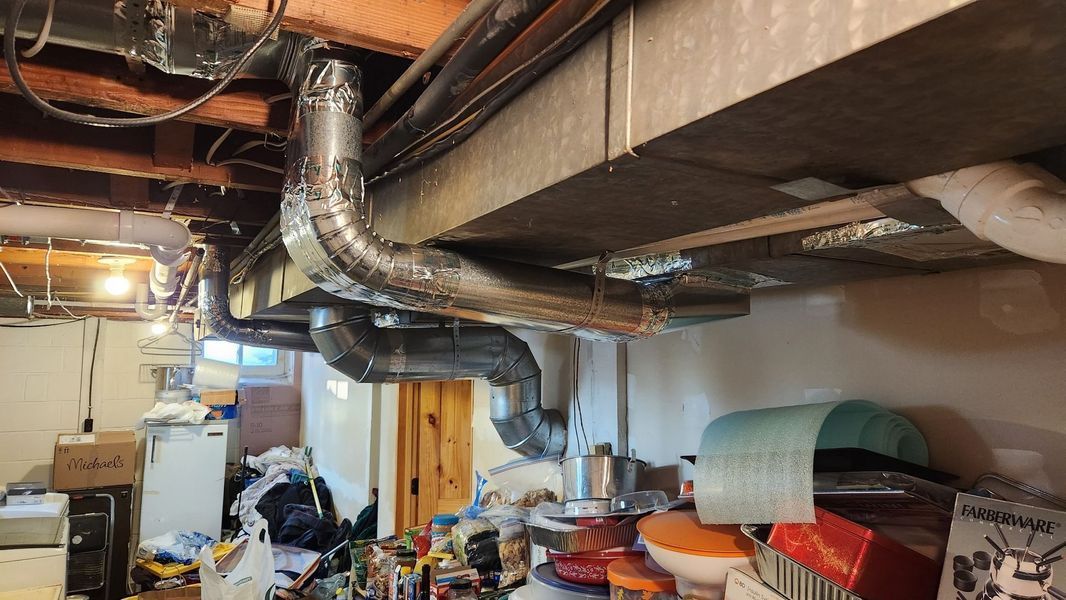 Basement with ductwork and cluttered storage. Pipes run along a wooden beam, with boxes and items on the floor.