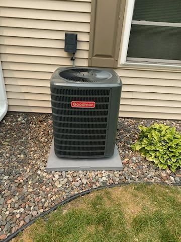 Goodman air conditioner unit on a concrete pad, surrounded by rocks and grass, near a house.
