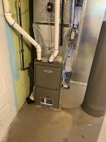 Furnace in a basement.  Gray metal unit with white PVC pipes, near a wall and a large metal duct.