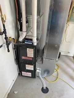 Furnace unit with vent pipes and ductwork in a utility room. Grey metal and white plastic visible.
