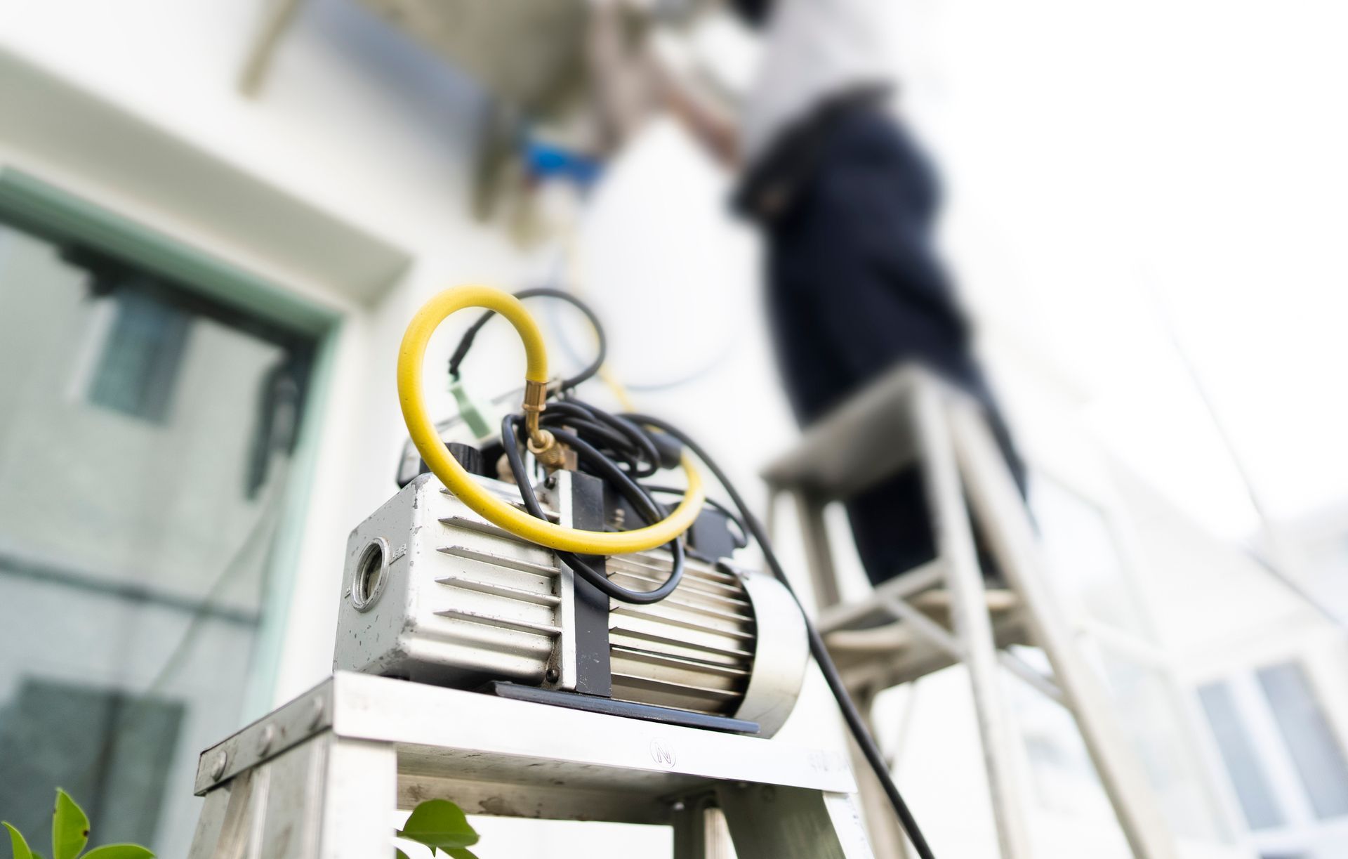 Vacuum pump on a ladder, connected to an AC unit being serviced by a technician outdoors.