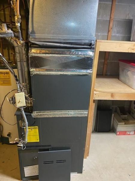 Furnace in a basement. The unit is grey and black with exposed ductwork, next to a wooden shelf.