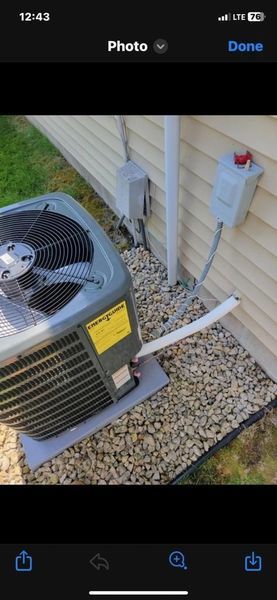 An air conditioning unit outside a building with electrical boxes and a drainage pipe resting on gravel.