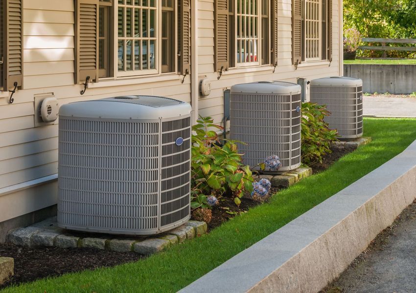 Three gray air conditioning units next to a light-colored building with green bushes and lawn.