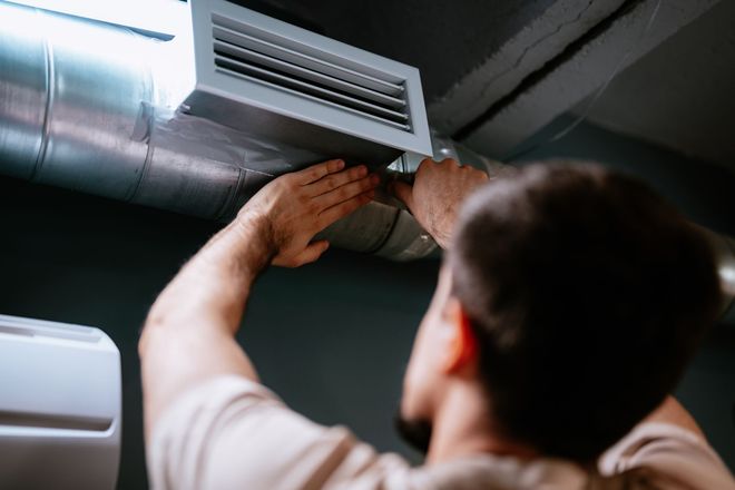 A person adjusting an air vent connected to silver ductwork on a dark ceiling.