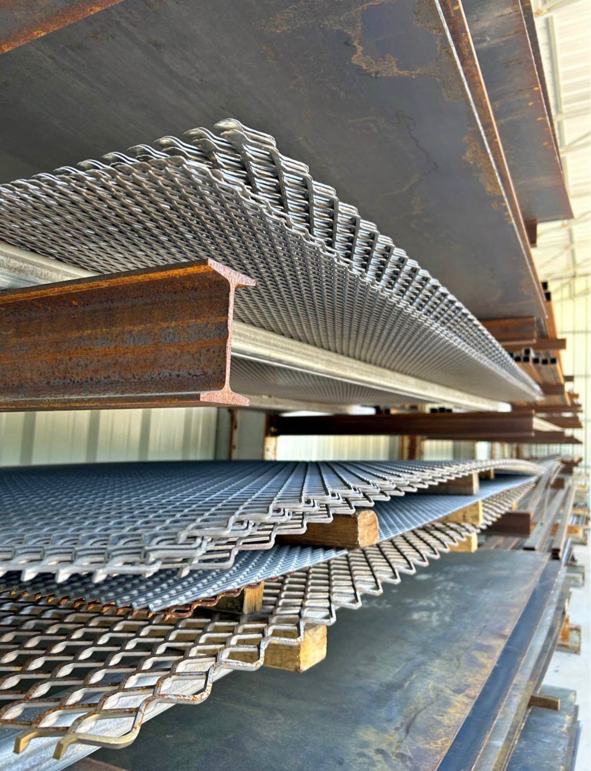 Metal sheets, various patterns, stacked on industrial shelves. Steel beams support the shelves.