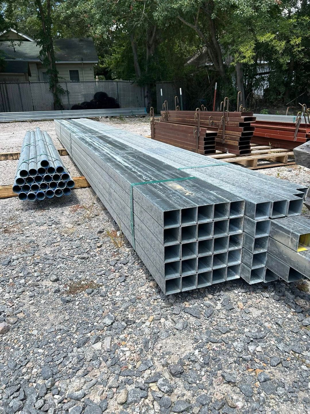 Stacks of metal square and round tubing outdoors on gravel.
