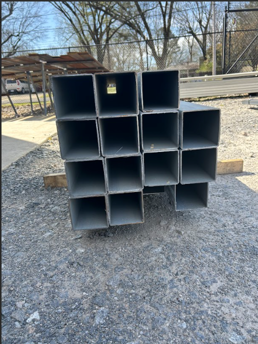 Stacked, square metal tubing on gravel, possibly construction materials, outdoors.