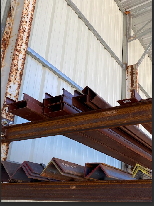 Steel beams and angle iron stacked on shelves in a warehouse, some with rust.