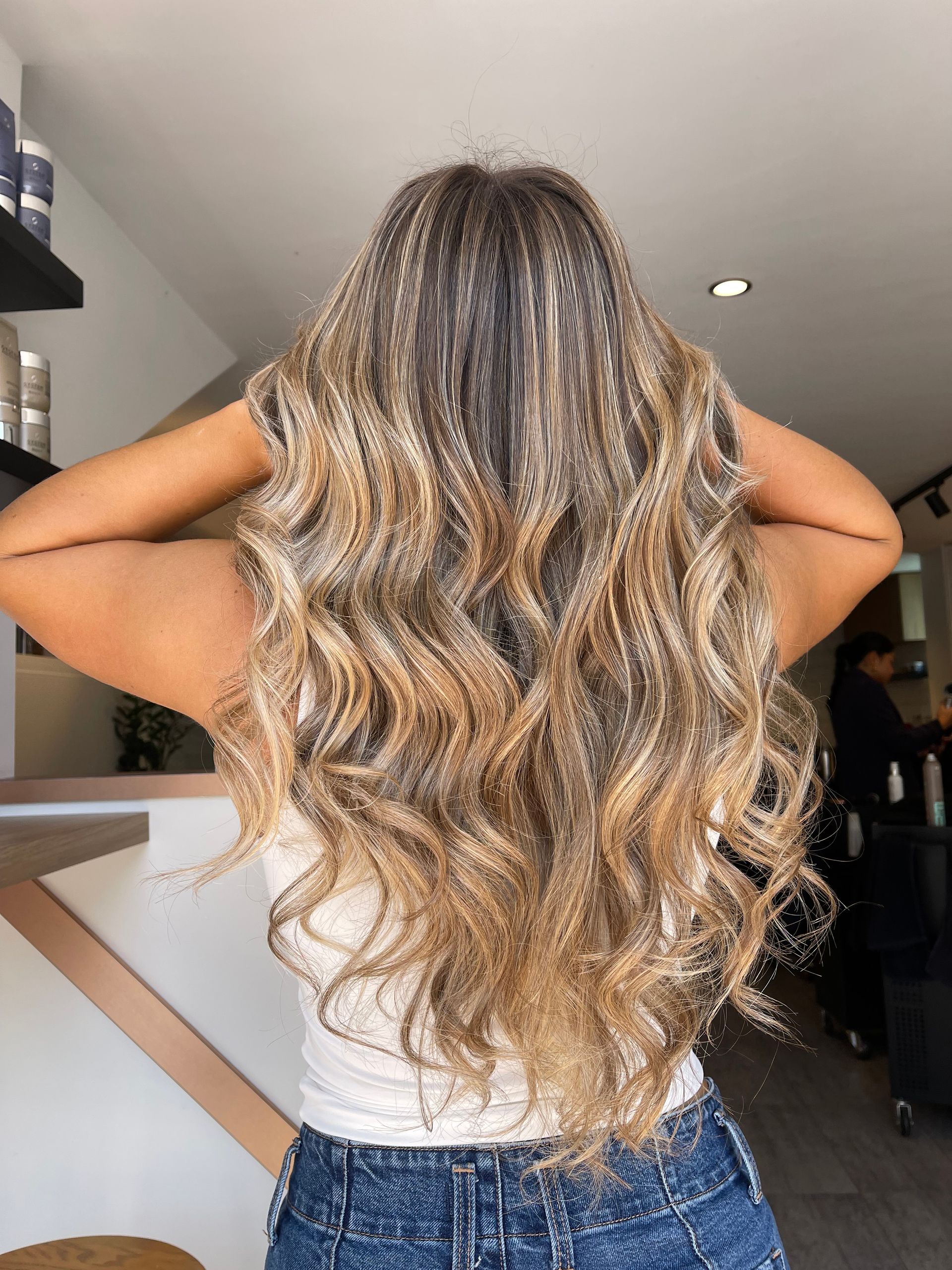 A woman with long blonde hair is standing in a salon with her hands behind her head.