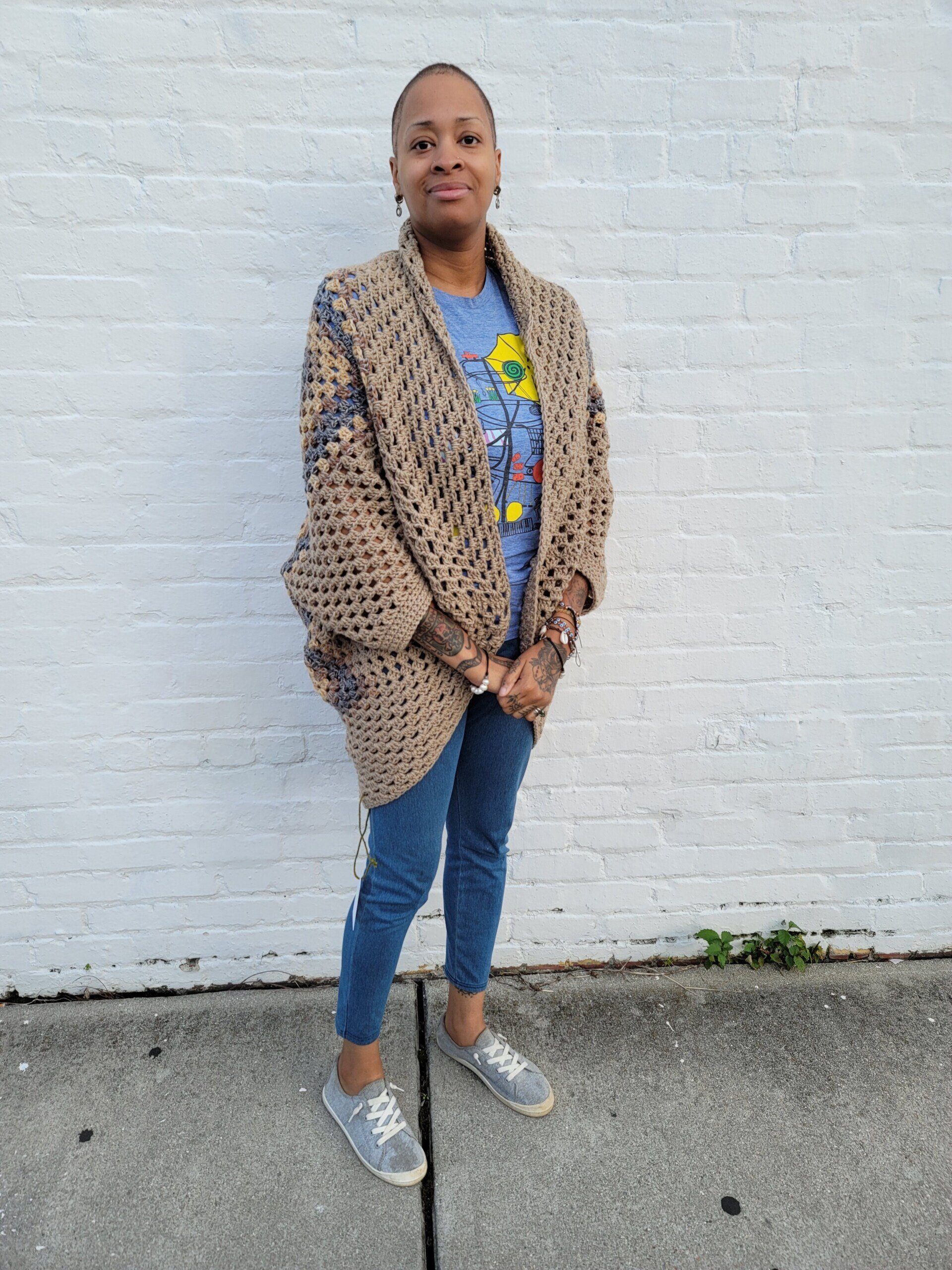 A woman wearing a crocheted cardigan is standing in front of a white brick wall.