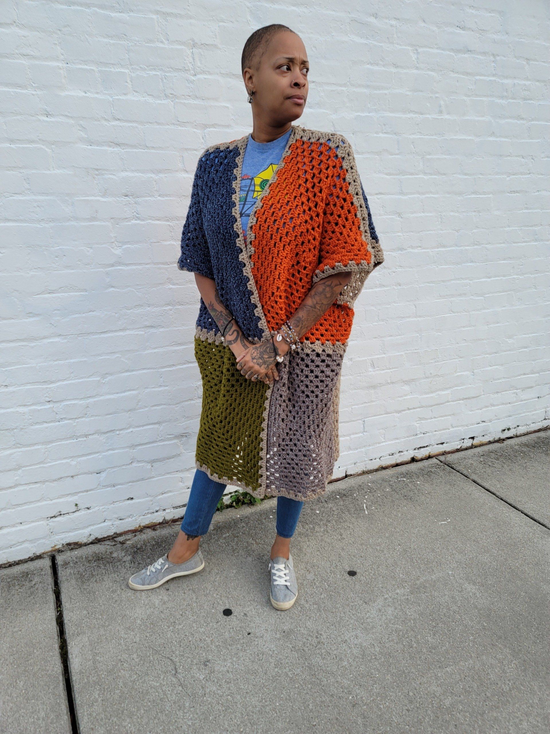 A woman wearing a crocheted cardigan is standing on a sidewalk in front of a white brick wall.