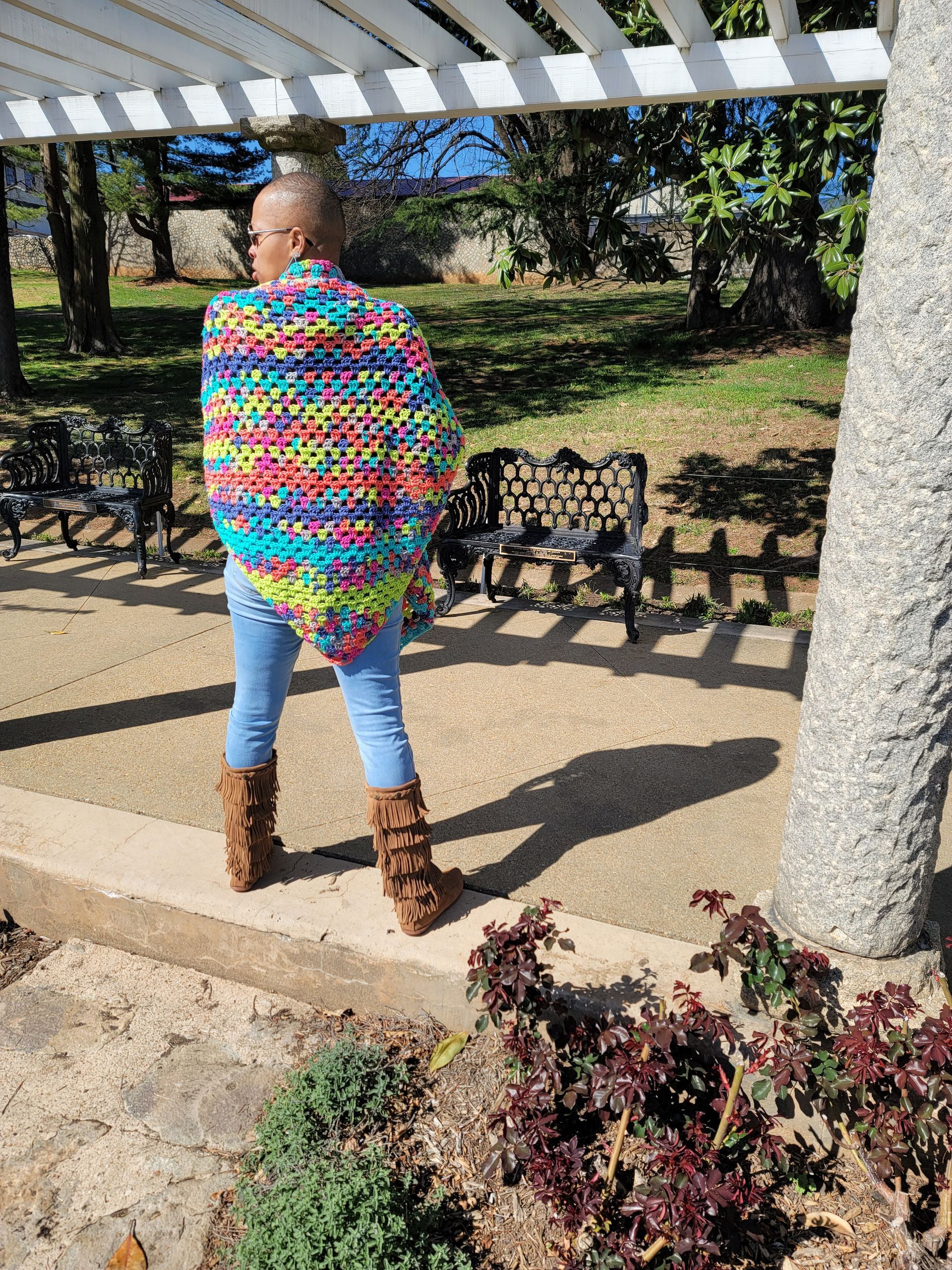 A woman wearing a colorful sweater and boots is standing under a pergola in a park.