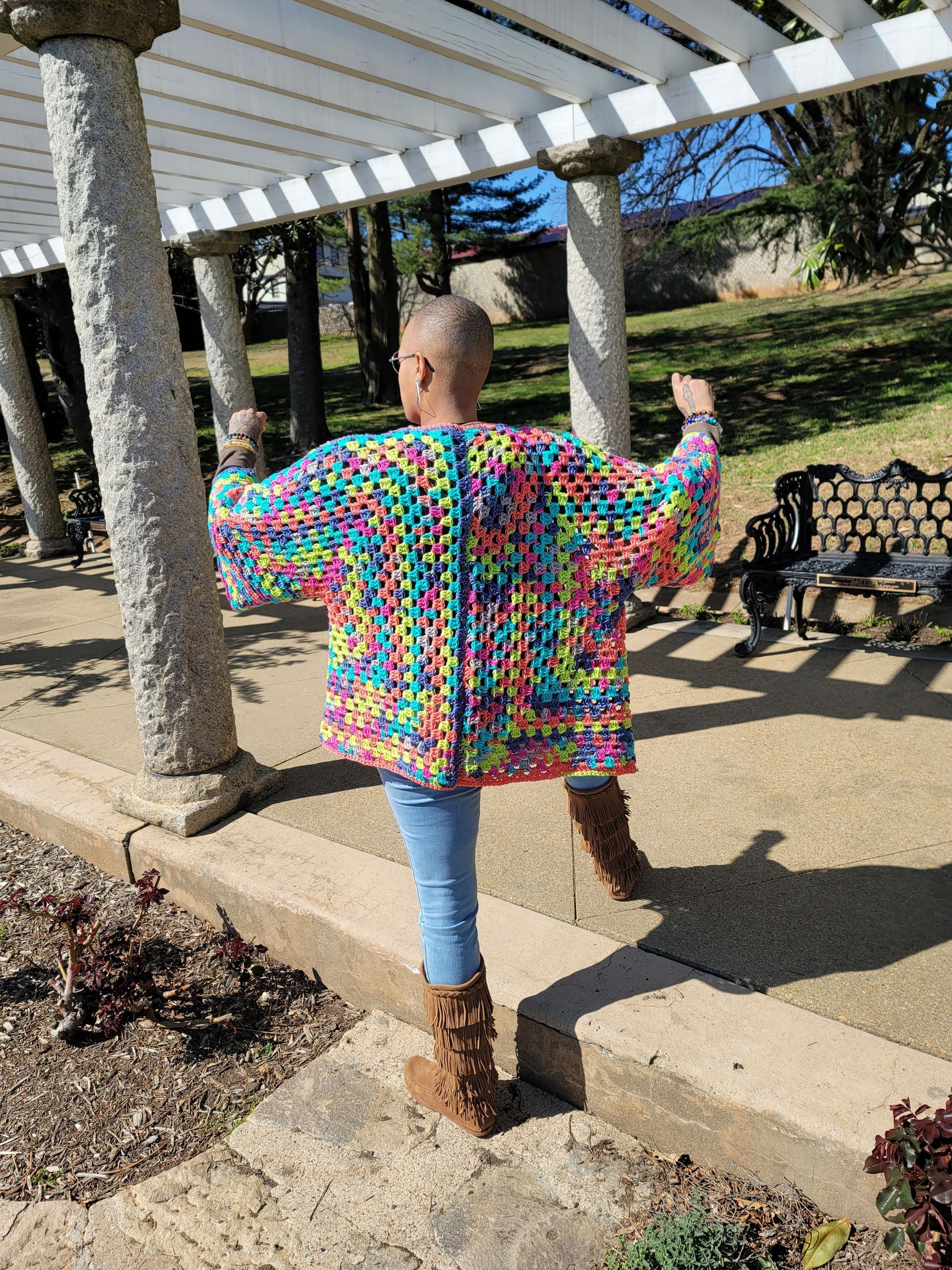 A woman wearing a colorful crocheted cardigan is standing in a park.