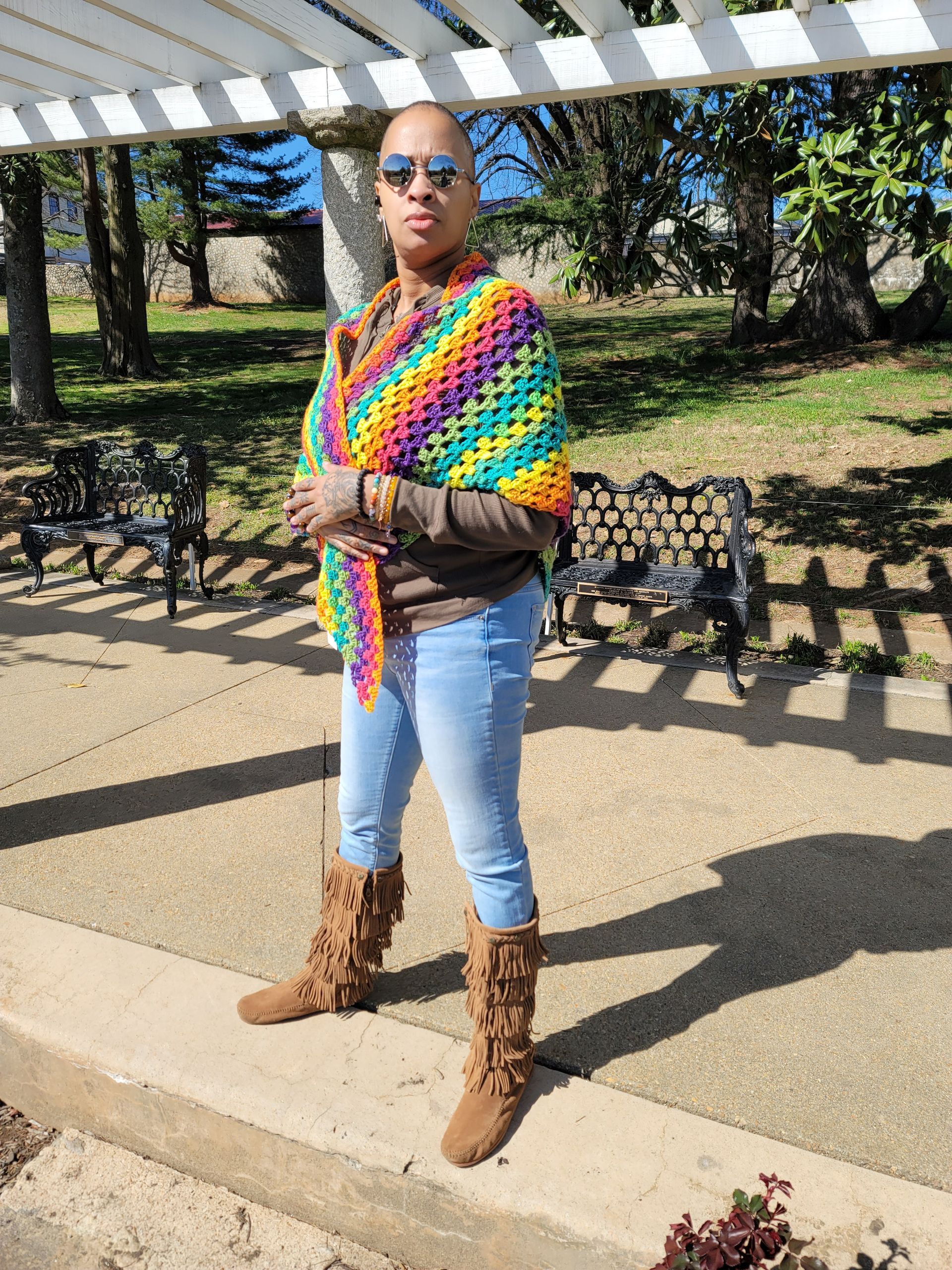 A woman wearing a crocheted shawl and boots is standing on a sidewalk in a park.