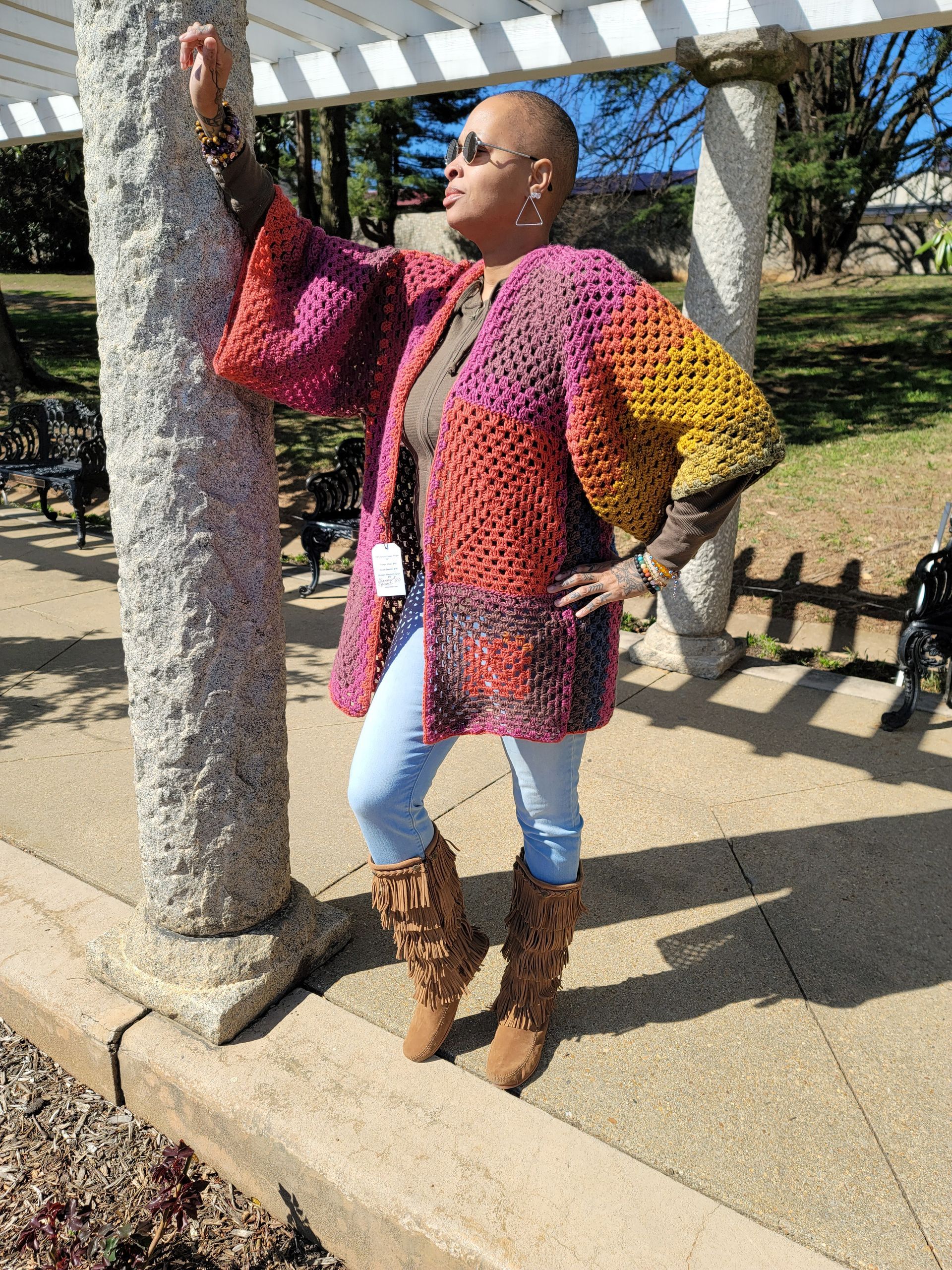 A woman wearing a crocheted cardigan and boots is standing next to a stone column.