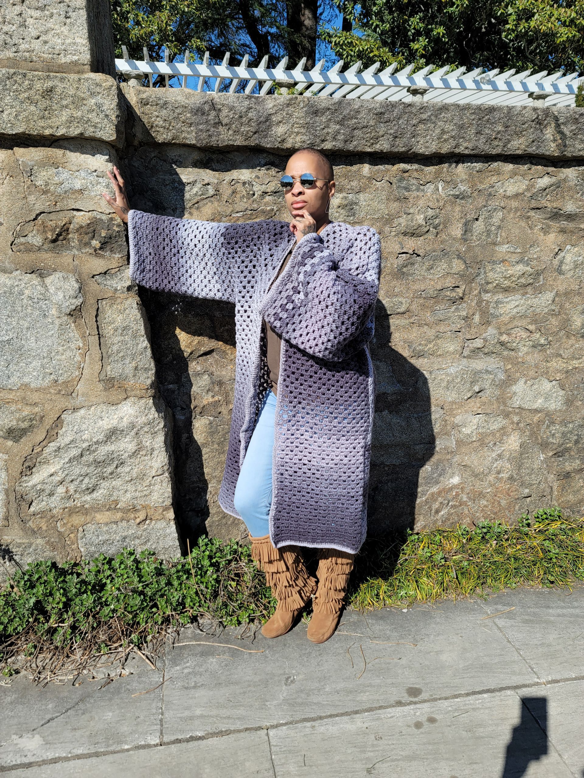 A woman is standing in front of a stone wall wearing a crocheted coat.