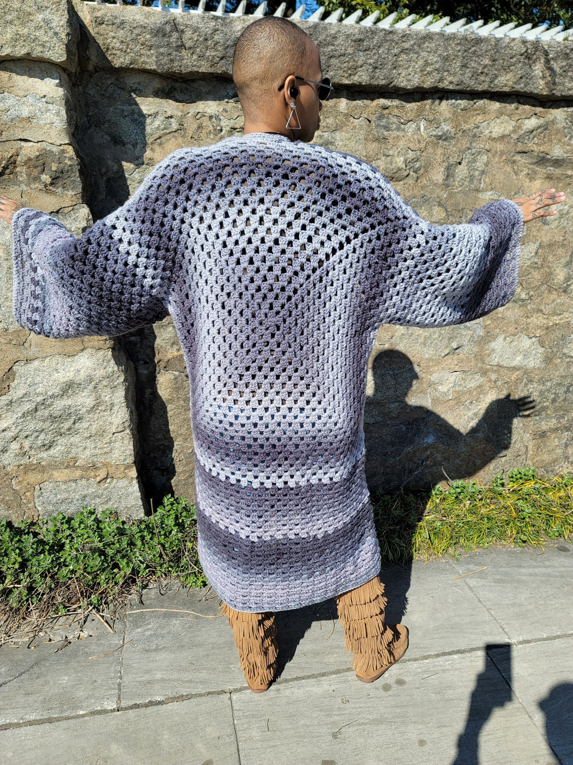A woman is standing on a sidewalk wearing a crocheted cardigan.