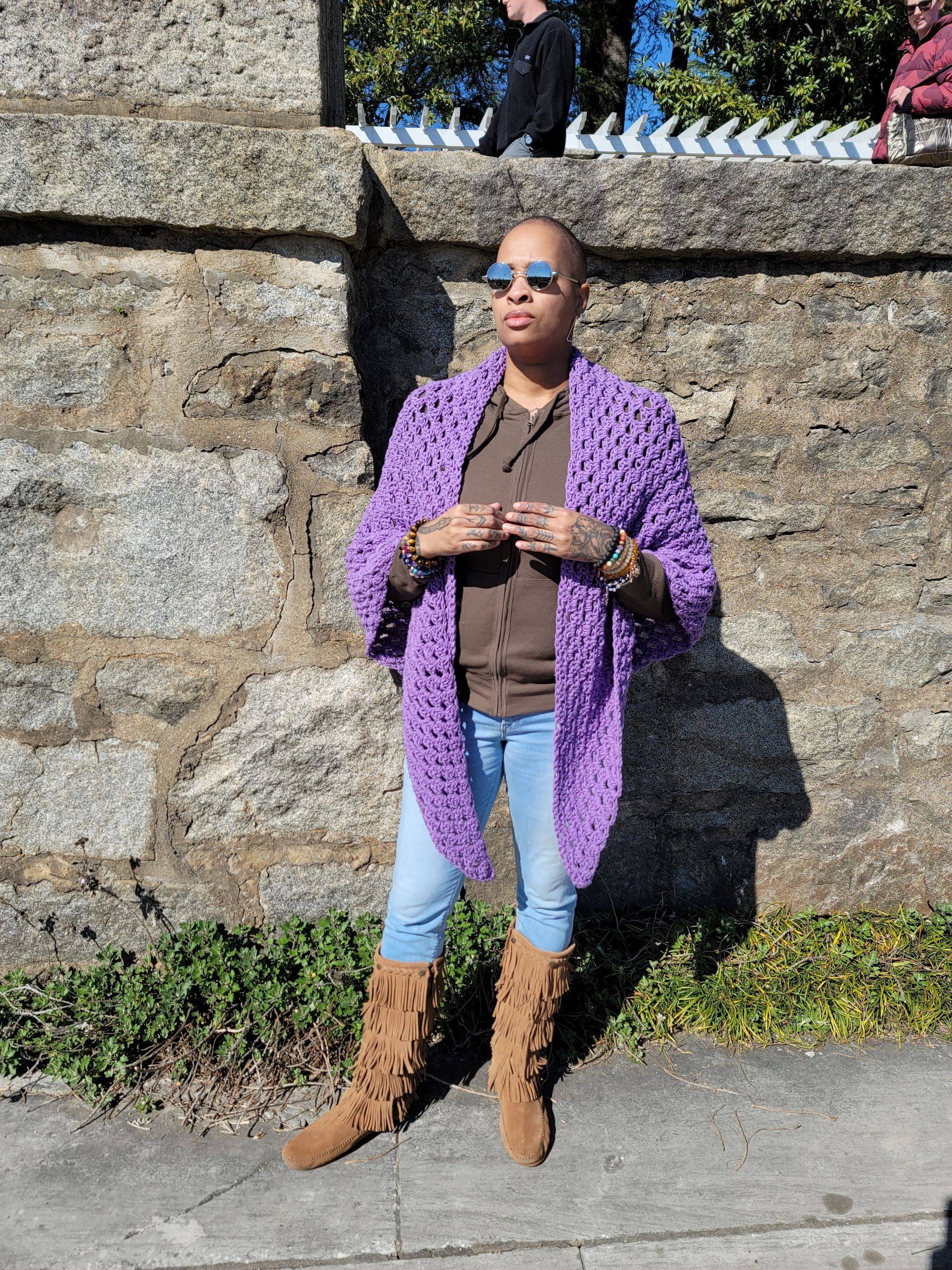 A woman wearing a purple cardigan and boots is standing in front of a stone wall.