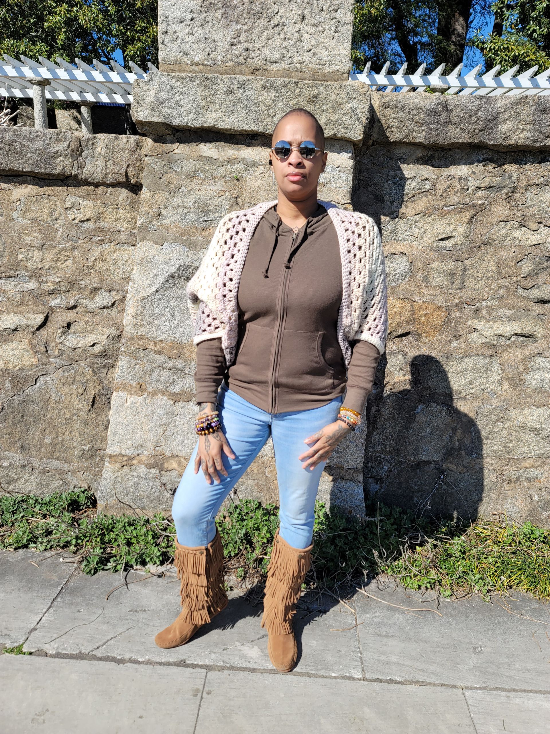 A woman wearing a sweater and boots is standing in front of a stone wall.