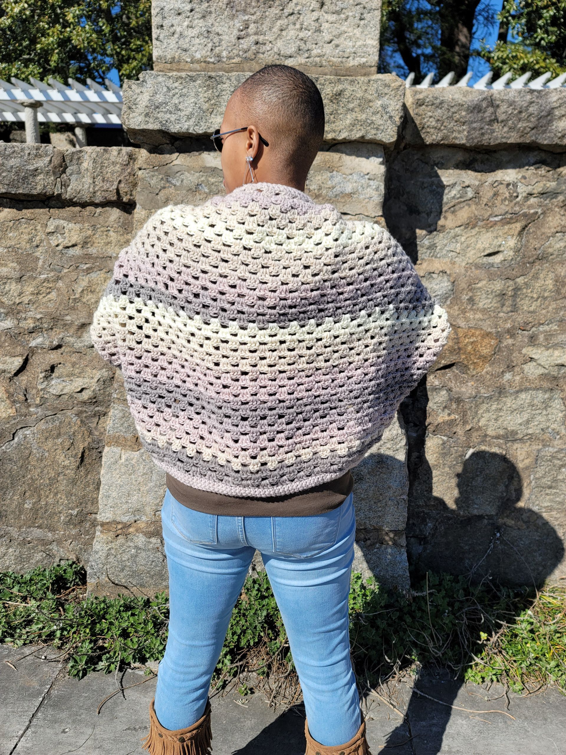 A woman wearing a crocheted sweater and jeans is standing in front of a stone wall.