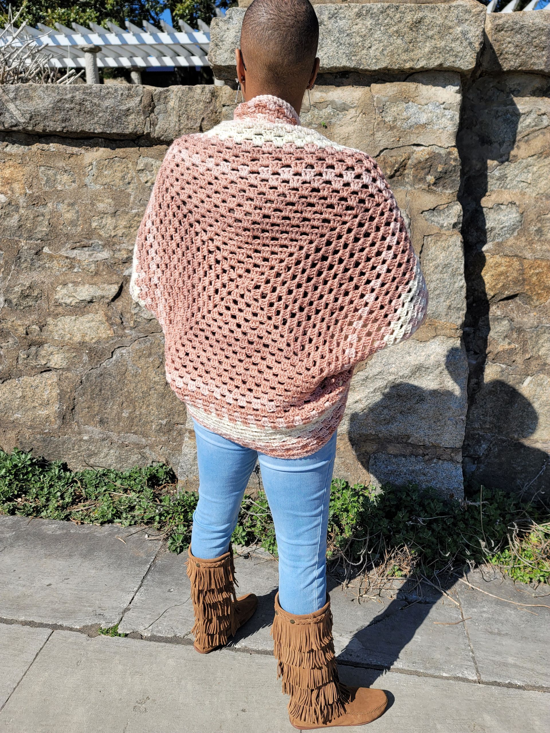 A woman wearing a crocheted shawl and boots is standing in front of a stone wall.