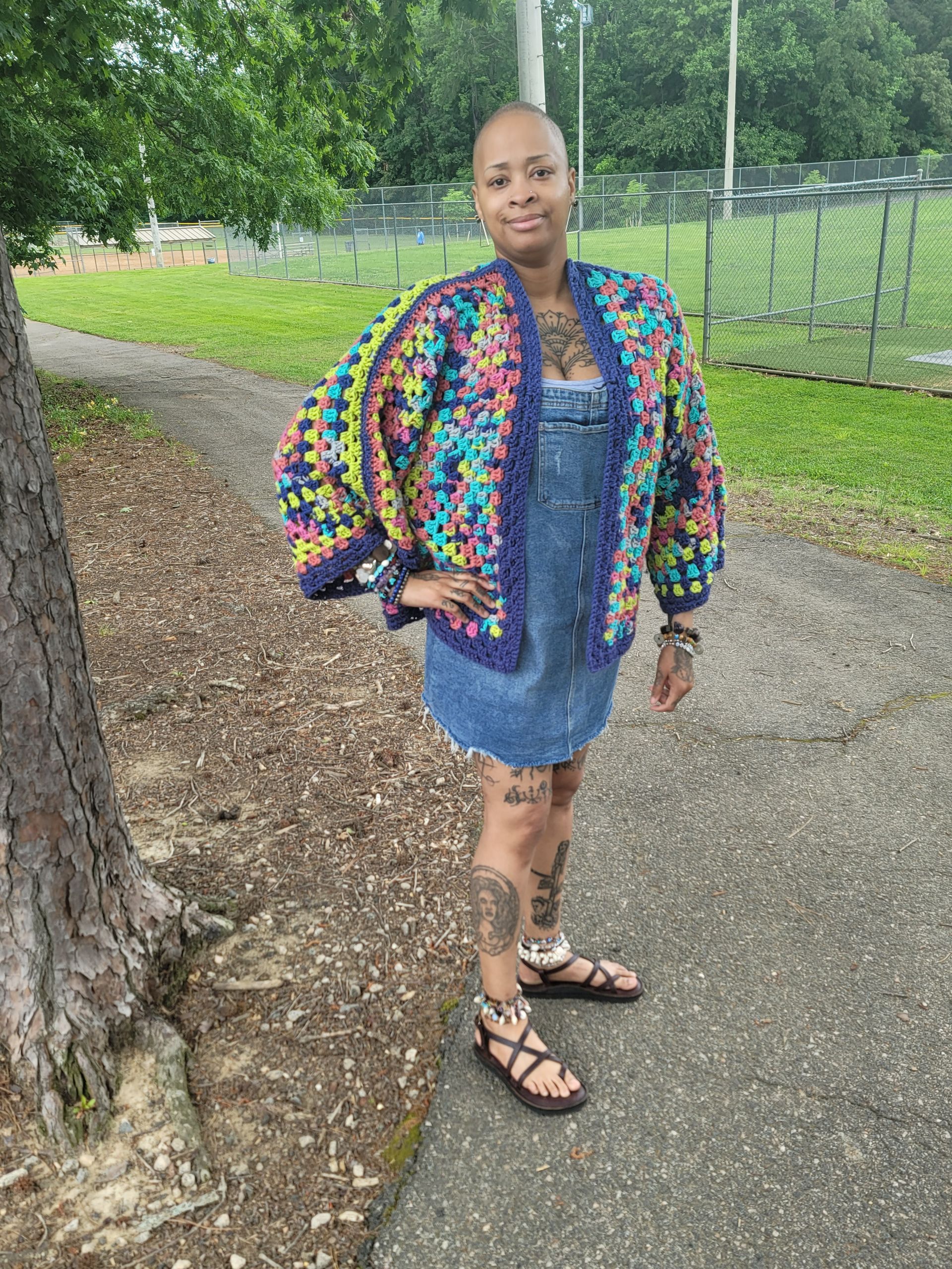 A woman in a crocheted jacket and denim dress is standing on a sidewalk.