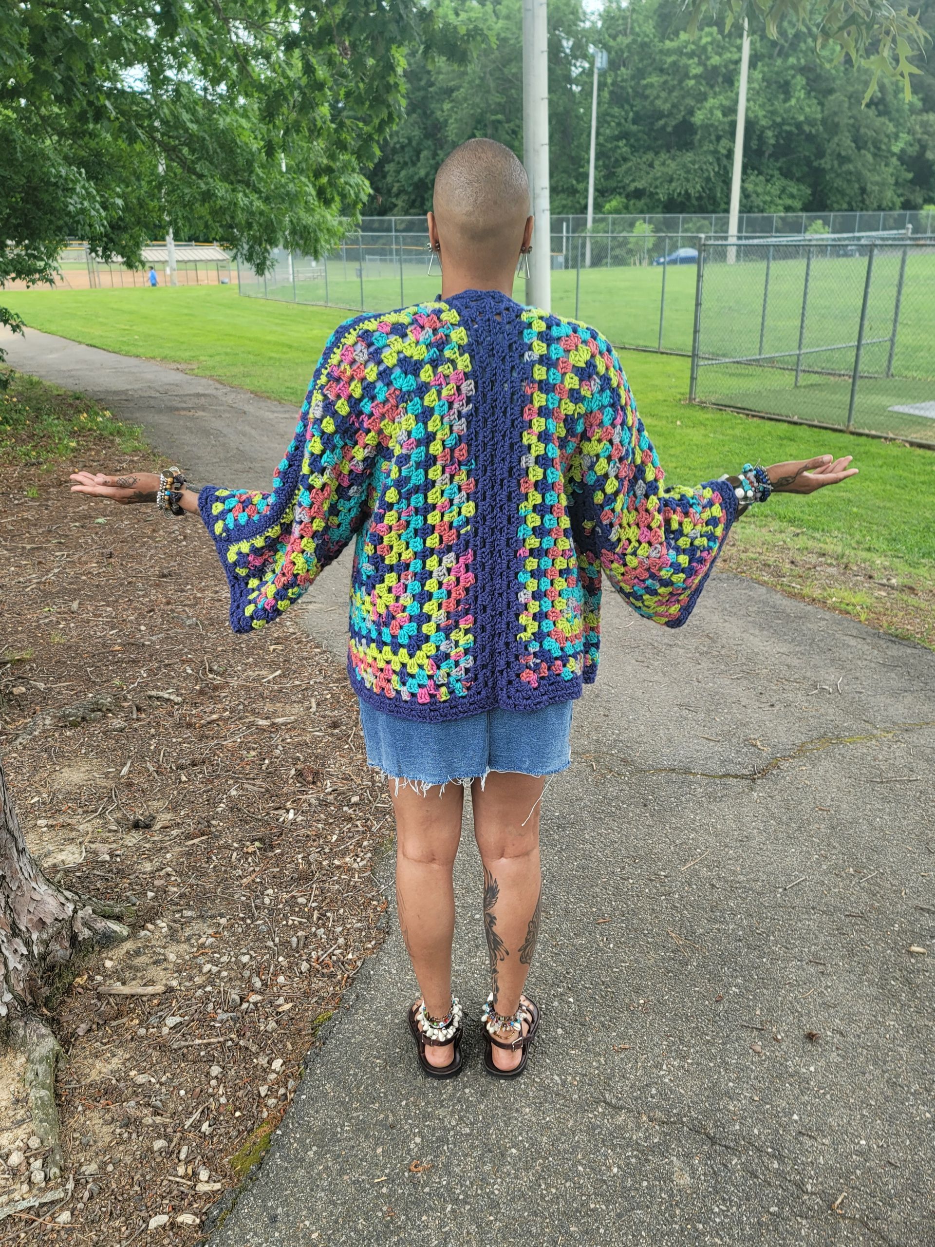 A woman wearing a colorful crocheted cardigan is standing on a sidewalk with her arms outstretched.