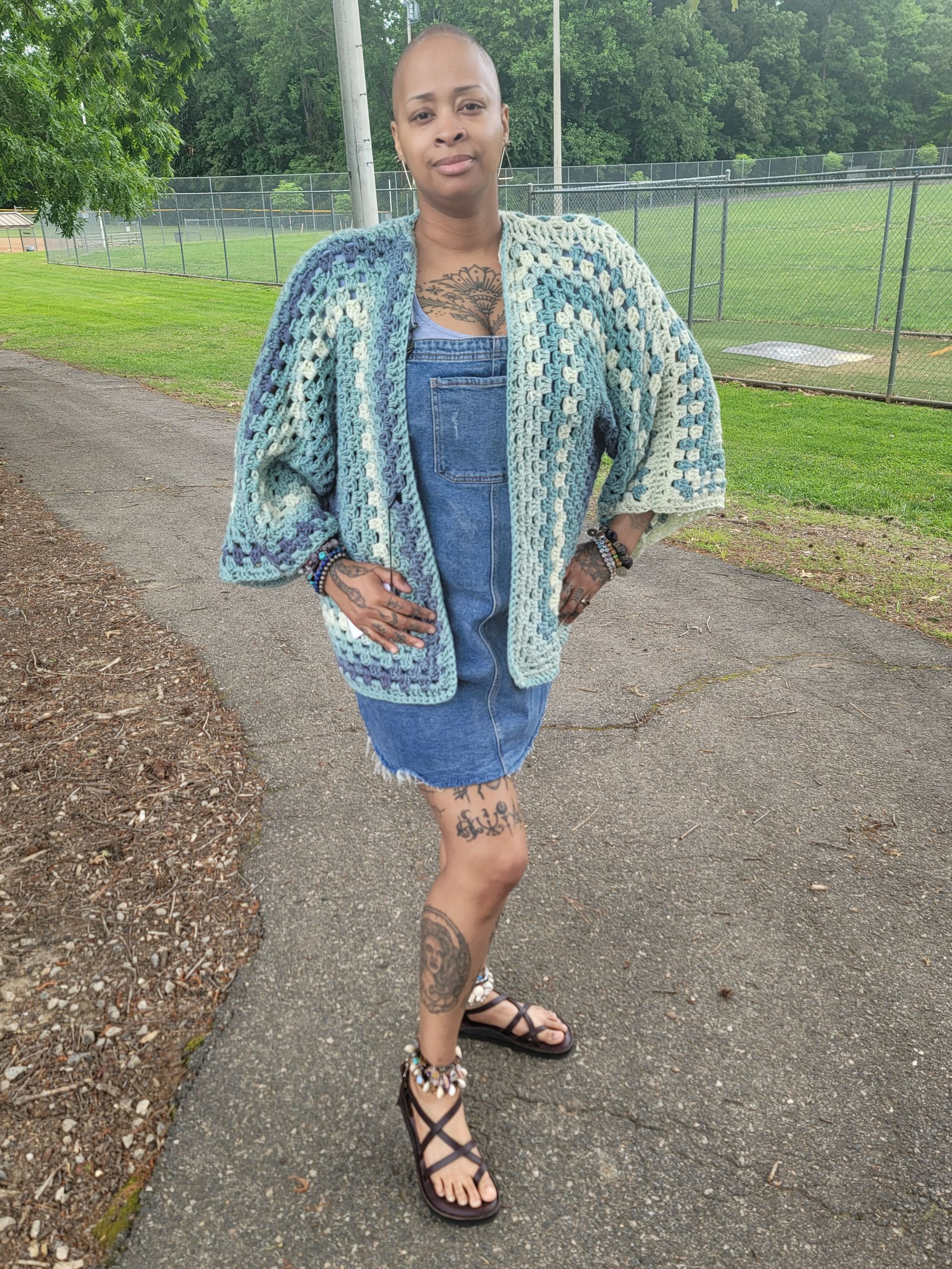 A woman wearing a crocheted jacket and overalls is standing on a sidewalk.