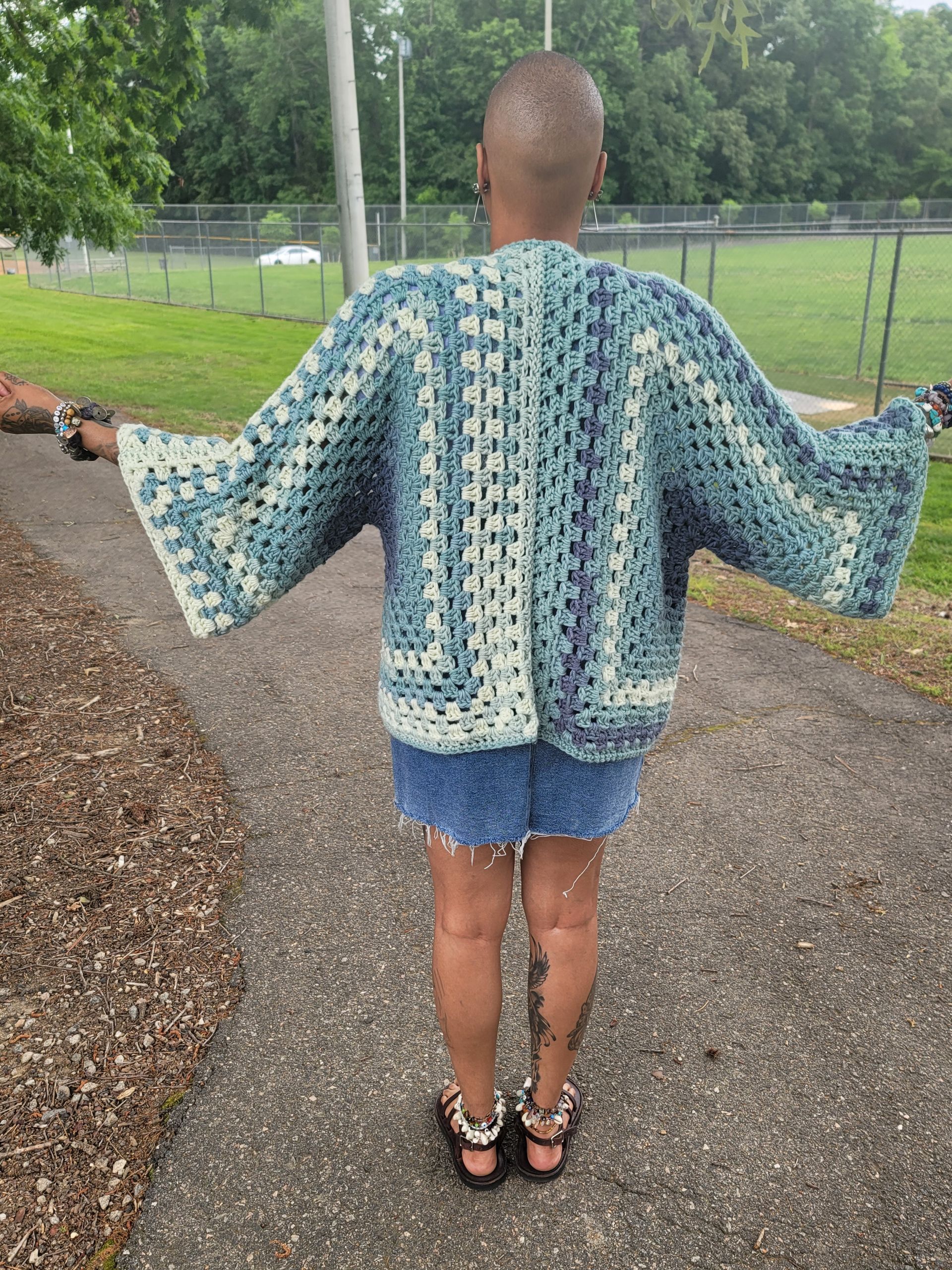 A woman is standing on a sidewalk wearing a crocheted jacket.