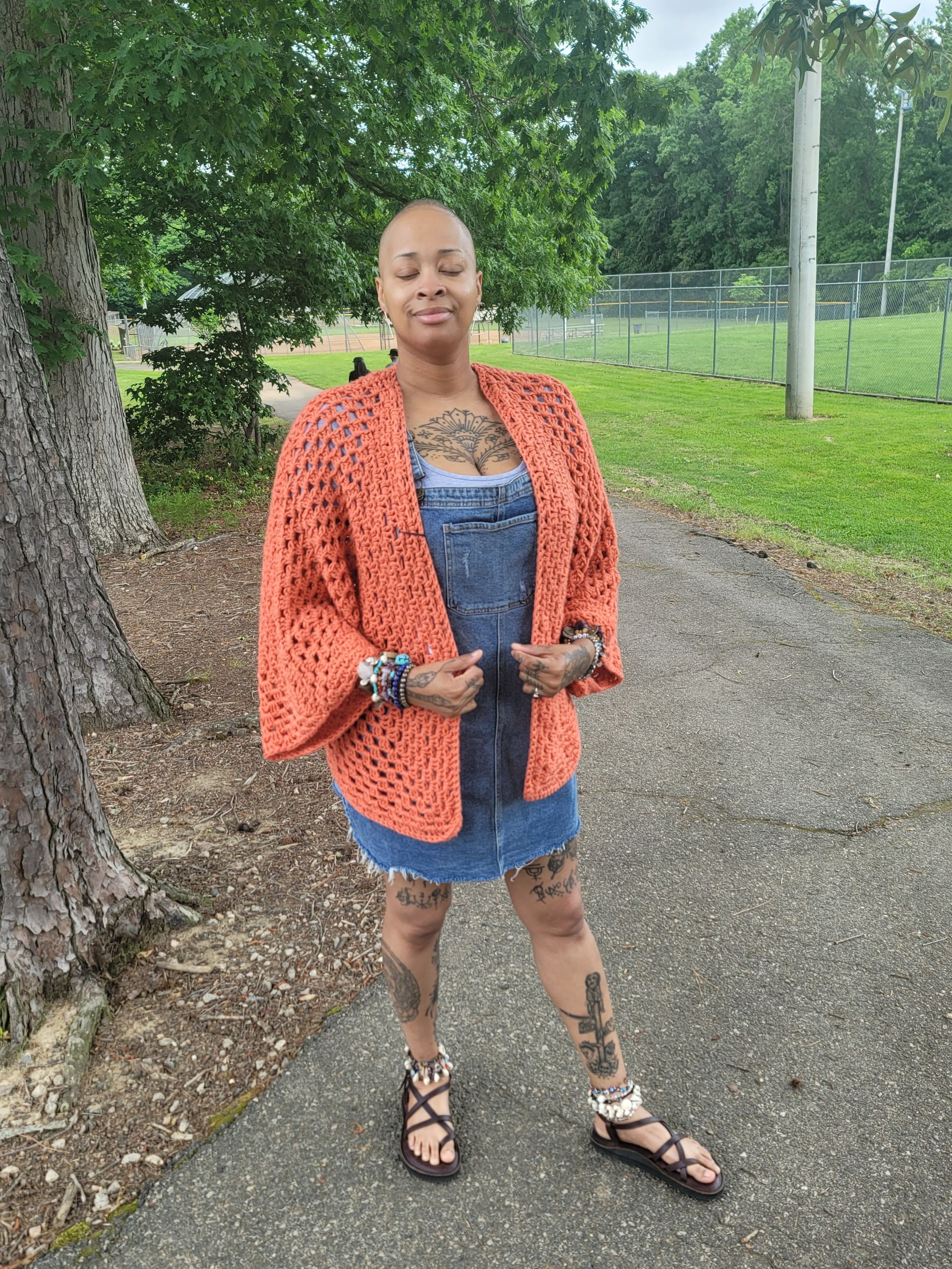 A woman is standing on a sidewalk wearing a crocheted cardigan and overalls.