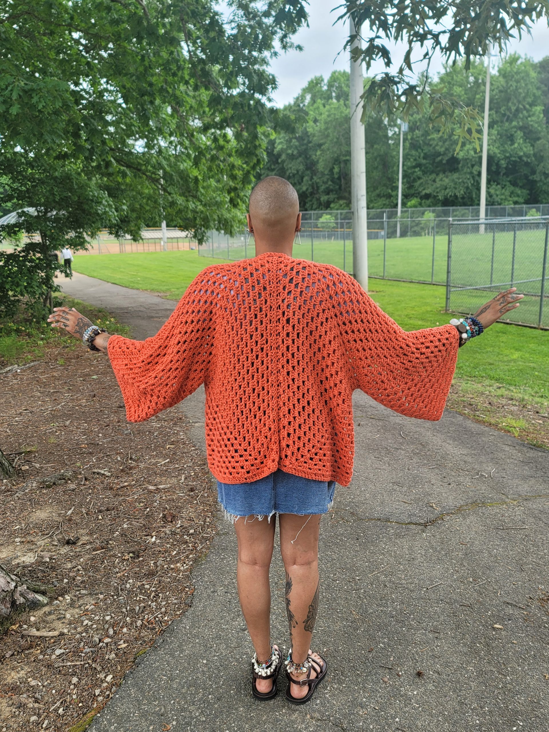 A woman wearing a crocheted cardigan and shorts is standing on a sidewalk with her arms outstretched.
