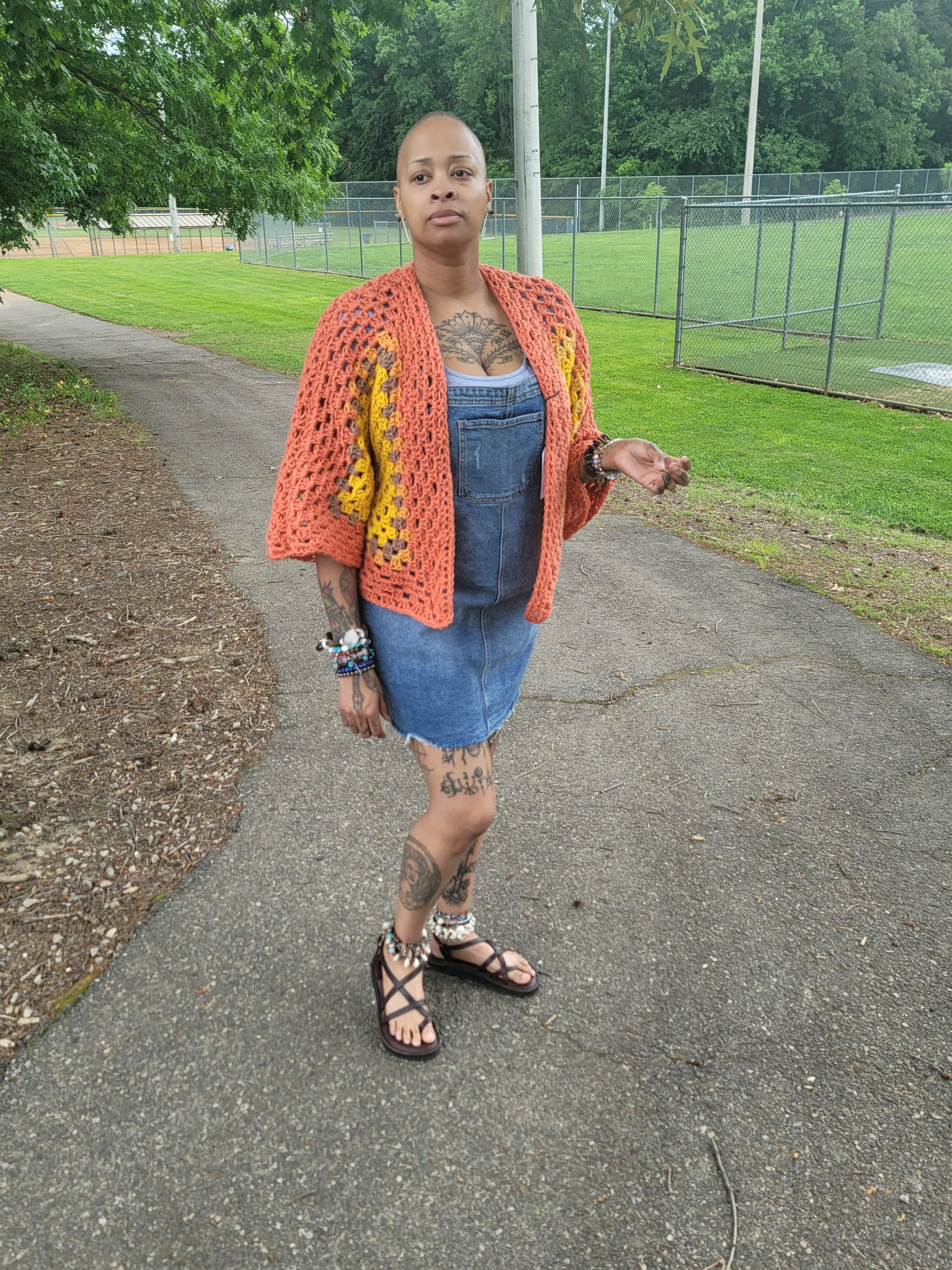 A woman is standing on a sidewalk wearing a crocheted cardigan and overalls.