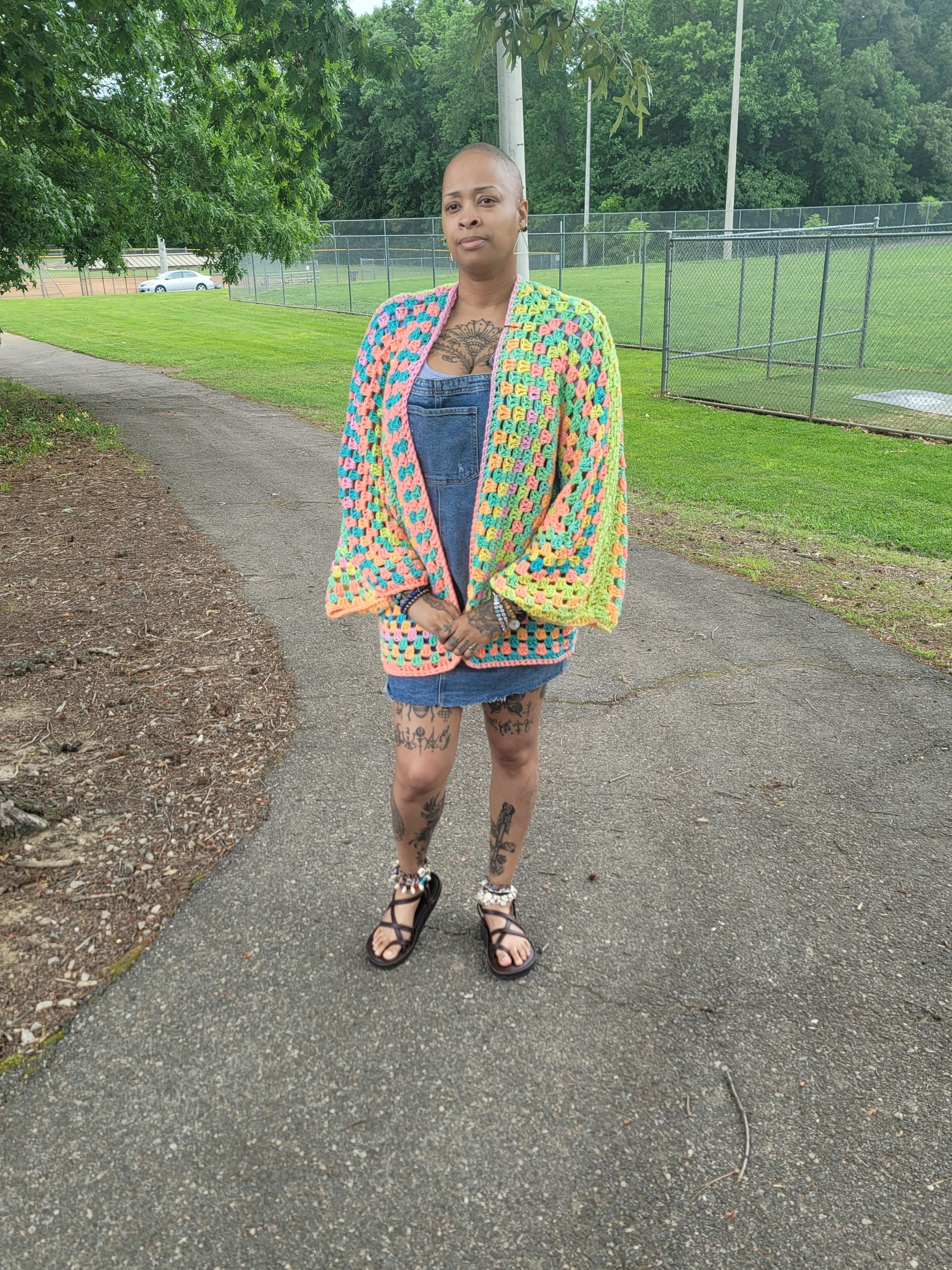 A woman is standing on a sidewalk wearing a colorful crocheted jacket.