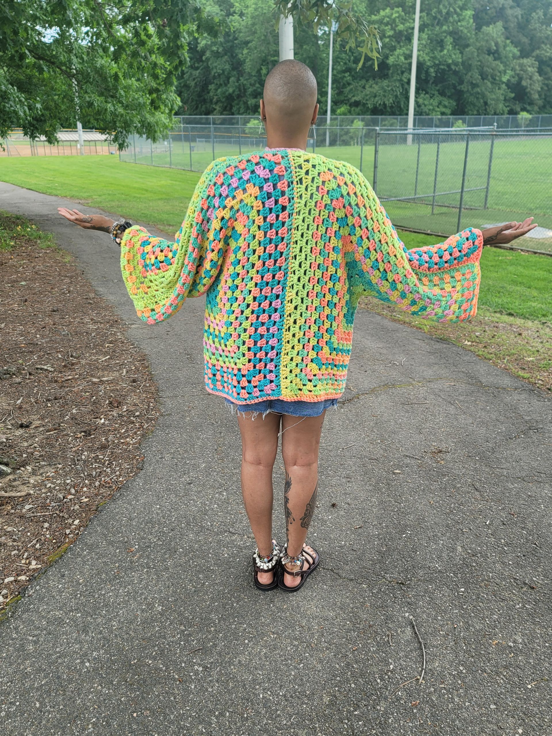 A woman is standing on a sidewalk wearing a colorful crocheted jacket.