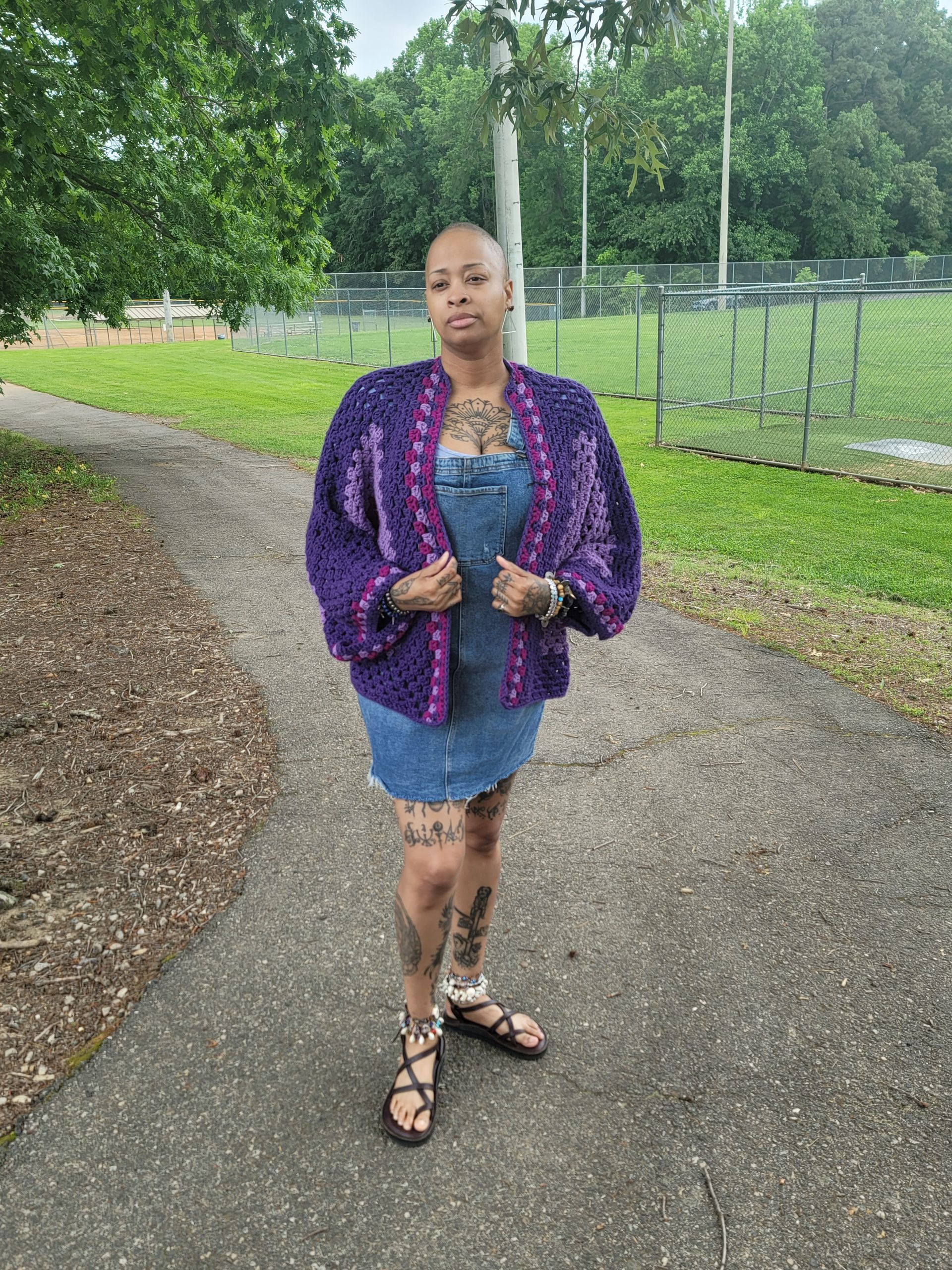 A woman wearing a purple cardigan and a denim dress is standing on a sidewalk.